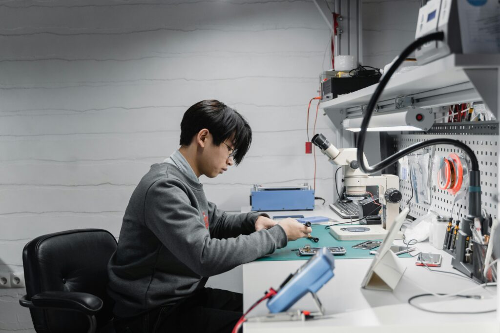 A young male engineer repairs electronics at a workshop desk with tools and equipment.